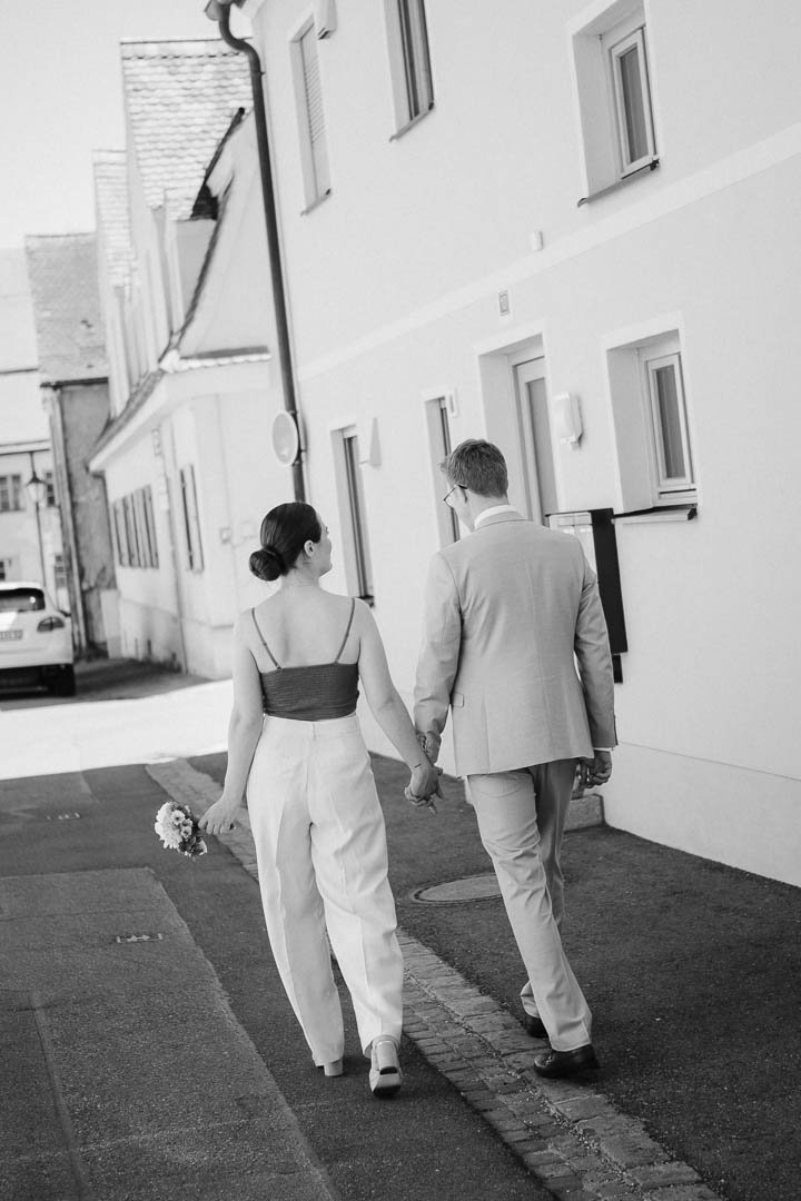 schräg und in schwarz-weiss aufgenommenes Hochzeitsfoto eines Brautpaares, das von hinten zu sehen ist, während es Hand in Hand eine Strasse in der Altstadt von Augsburg entlang geht
