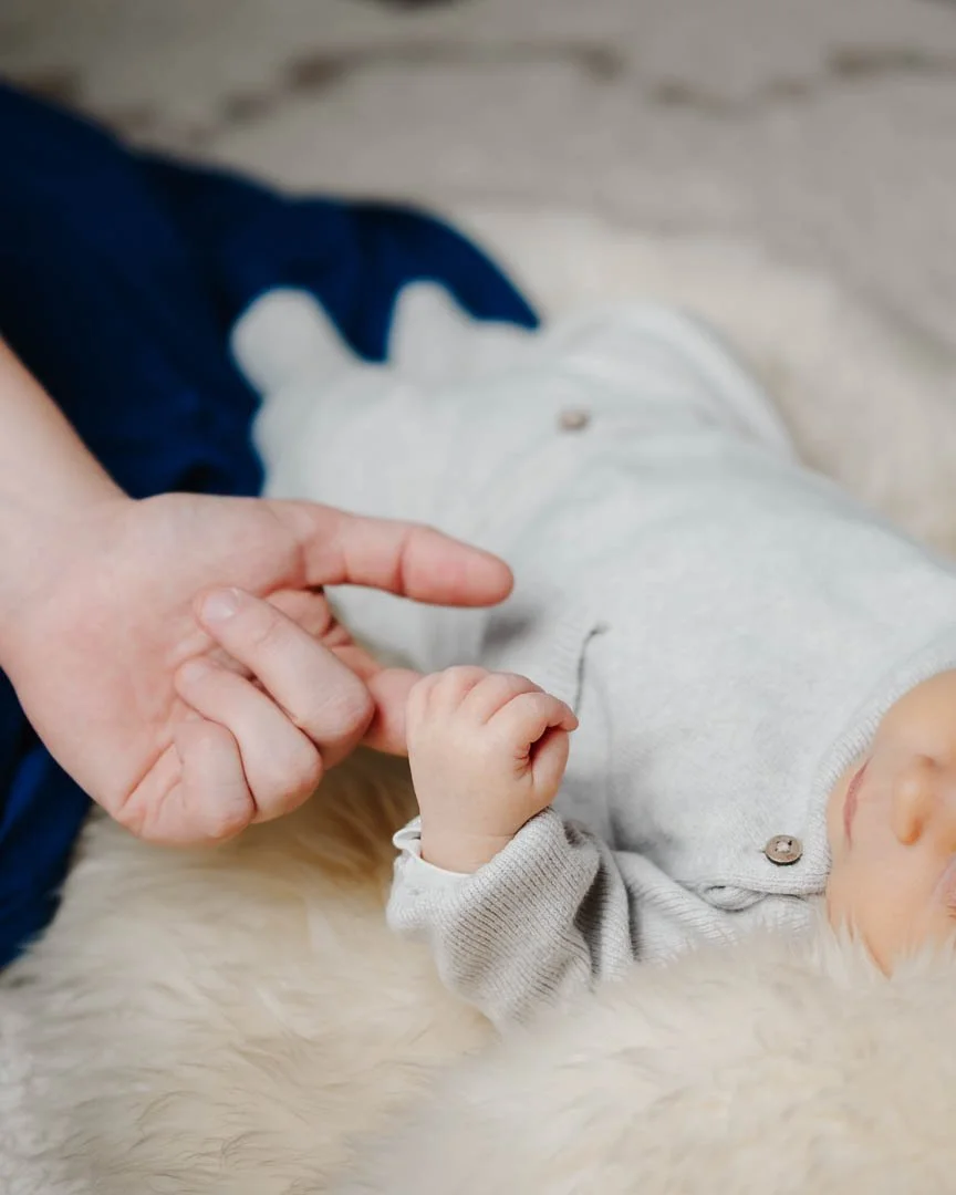 Newborn-Portrait, das ein Baby auf einem Fell liegend zeigt, welches mit seiner Hand den Finger seiner Mama umgreift
