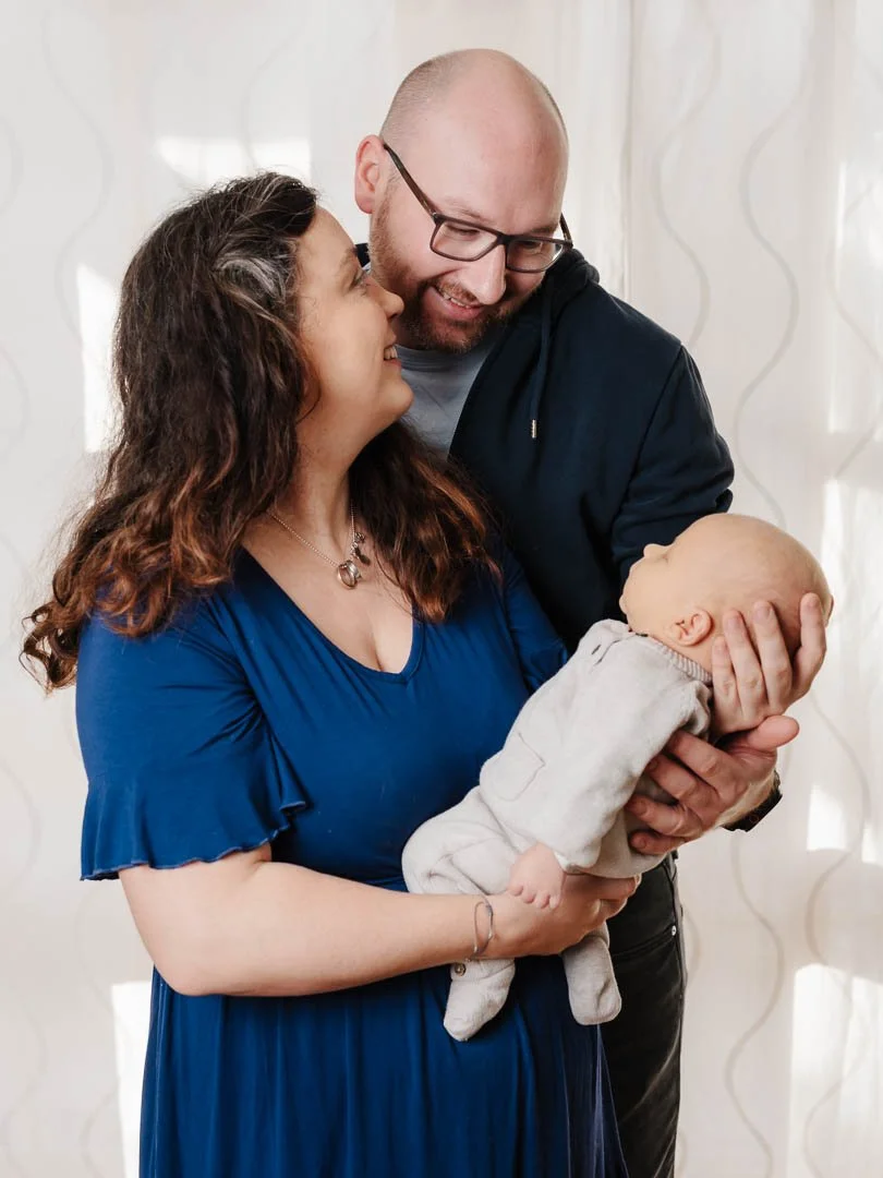 Newborn-Foto aus Augsburg, auf dem ein glückliches Paar in der eigenen Wohnung sein neugeborenes Baby auf dem Arm hält und sich glücklich anschaut