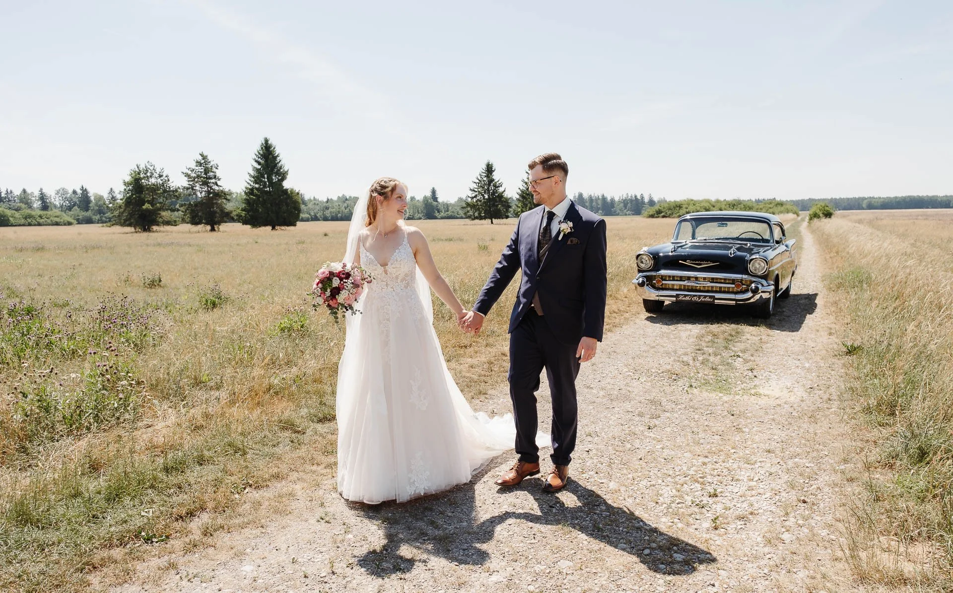 Hochzeitsfotografie, auf der ein Brautpaar mit klassischer Brautmode in einer sommerlichen Landschaft bei Augsburg spazieren geht, während im Hintergrund ein Oldtimer als Hochzeitsauto zu sehen ist