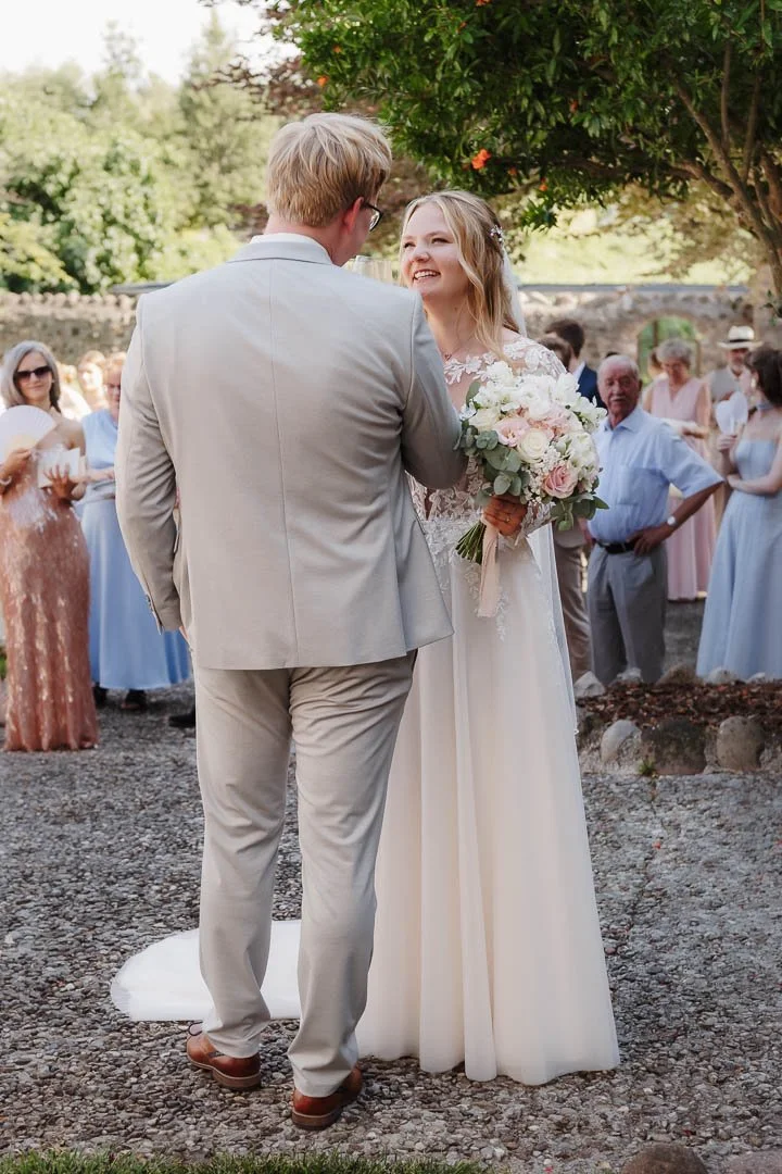 Foto eines Paares, das sich auf ihrer Hochzeit in Italien gegenübersteht und glücklich in die Augen blickt, während im Hintergrund unscharf die Hochzeitsgäste in pastellfarbenen Kleidern zu sehen ist