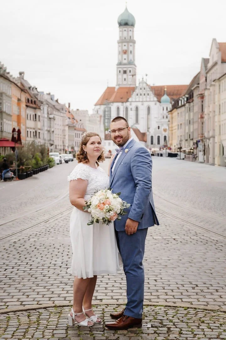 Ein Prautpaar steht nach der standesamtlichen Trauung in der Maxstraße in Augsburg eng beieinander, während im Hintergrund die Kirche St. Ulrich und historische Gebäude zu sehen sind