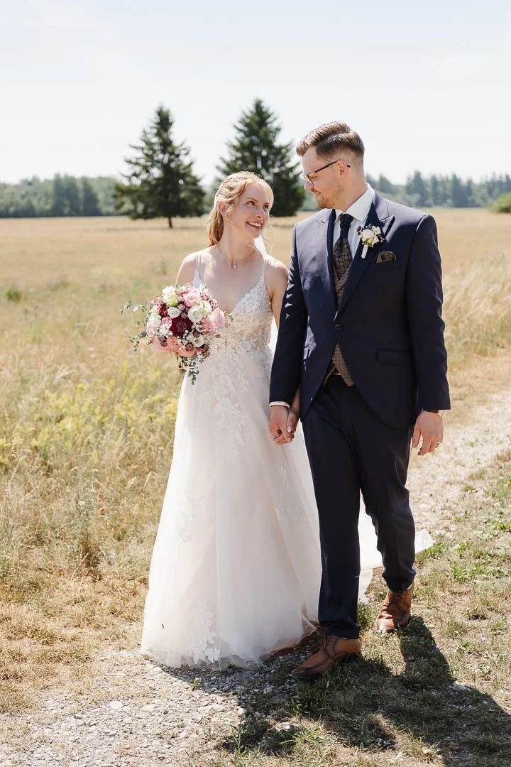 Hochzeitspaar mit klassischen Hochzeitsoutfits auf einem Feldweg in einer sonnigen Landschaft aus Wiesen und Wäldern bei Augsburg