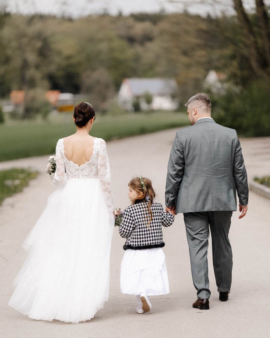 dokumentarisches Hochzeitsfoto eines Hochzeitspaares, das Hände haltend mit ihrem Kind eine Straße in der Region Stauden bei Augsburg entlang geht