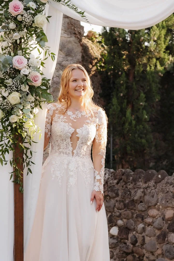 Portrait einer Braut, die im Außenbereich ihrer Hochzeitslocation am Gardasee neben Blumenschmuck steht und glücklich zur Seite blickt