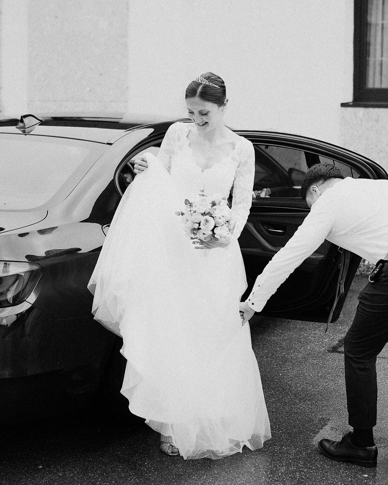 Schwarz-weiß Hochzeitsfoto, das eine Braut in Augsburg beim Aussteigen aus einem schwarzen Hochzeits-Auto zeigt, während sich der Trauzeuge um ihr Kleid kümmert