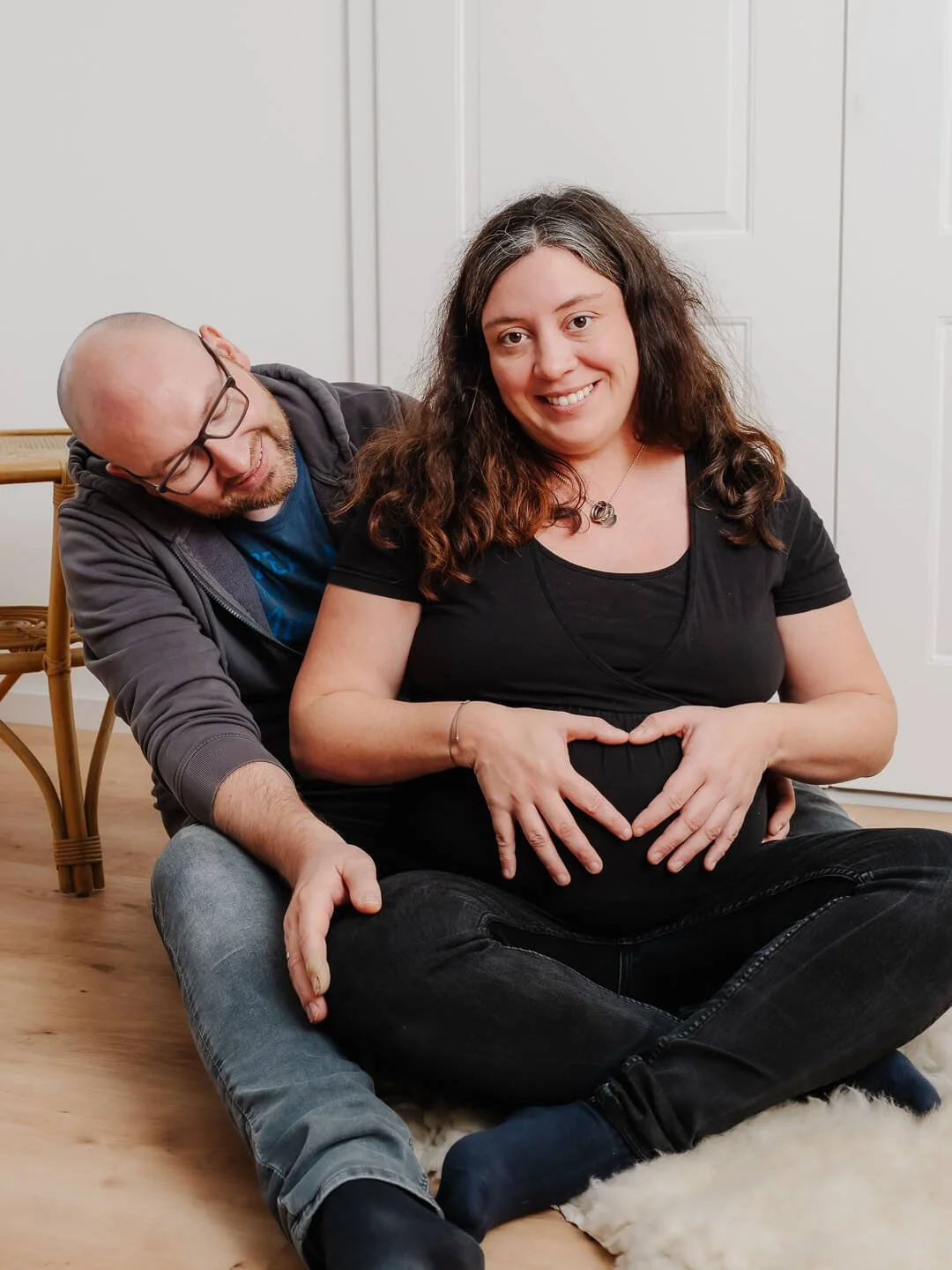 schwangere Frau mit ihrem Mann beim professionellen Babybauch-Fotoshooting auf dem Boden eines Wohnzimmers sitzend