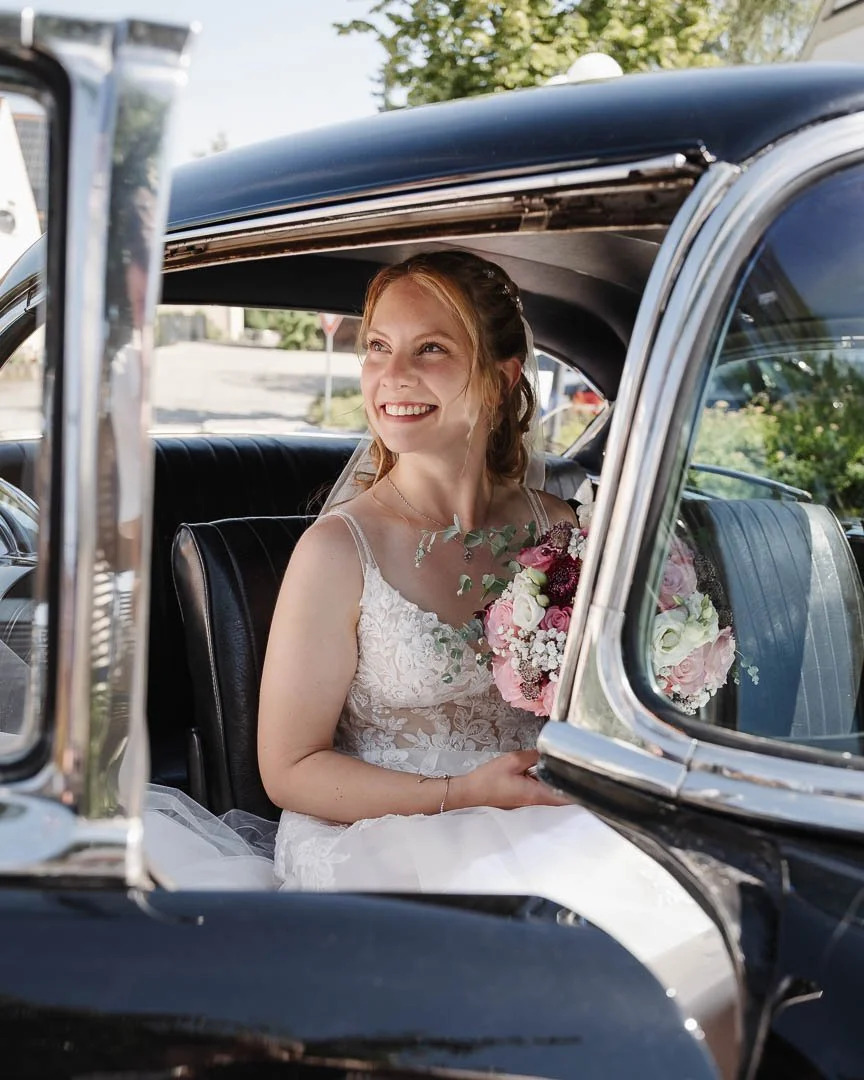 Portrait-Foto einer Braut, die glücklich lächelnd im Hochzeitsauto, einem Oldtimer aus Augsburg, sitzt