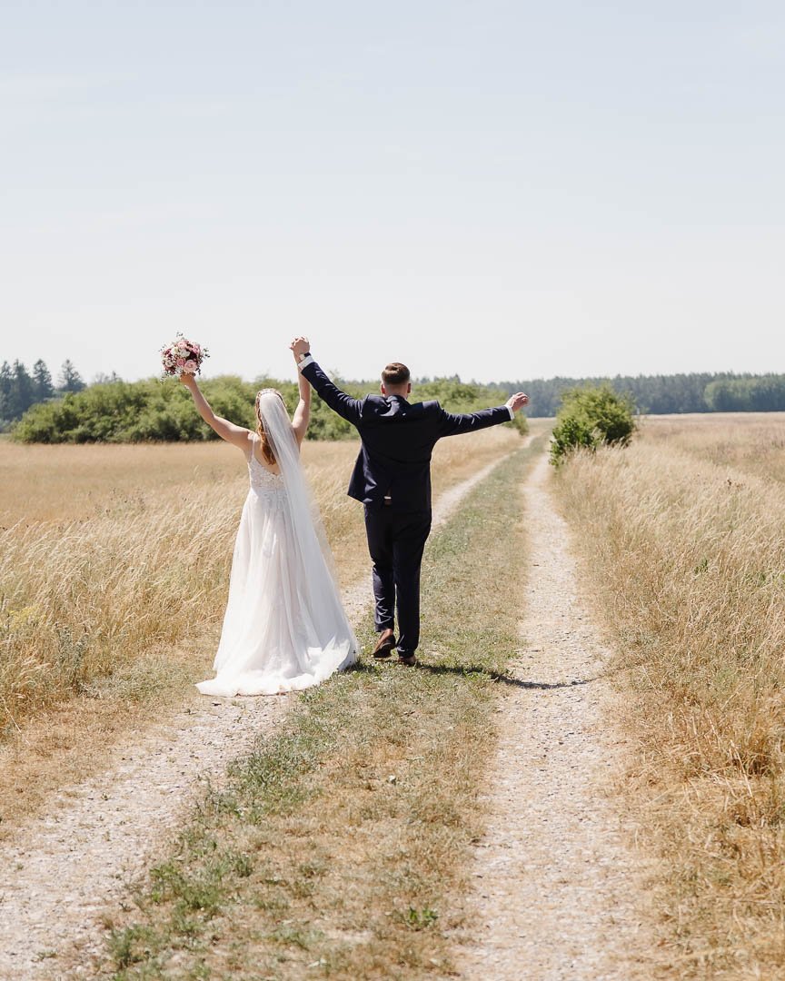 klassisches Hochzeitsfoto des Hochzeitsfotografen Philipp Tomaschko, das ein Brautpaar von hinten in der sommerlichen Landschaft um Augsburg zeigt, wie es einen Feldweg entlang geht und vor Freude die Hände in den Himmel wirft