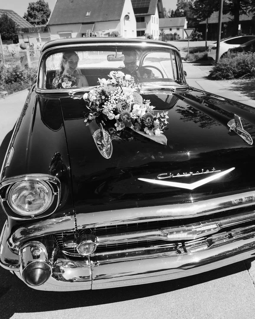 Das schwarz-weiß Bild zeigt ein Hochzeitsauto, einen Chevrolet Bel Air mit Blumenschmuck, in dem ein Hochzeitspaar sitzt
