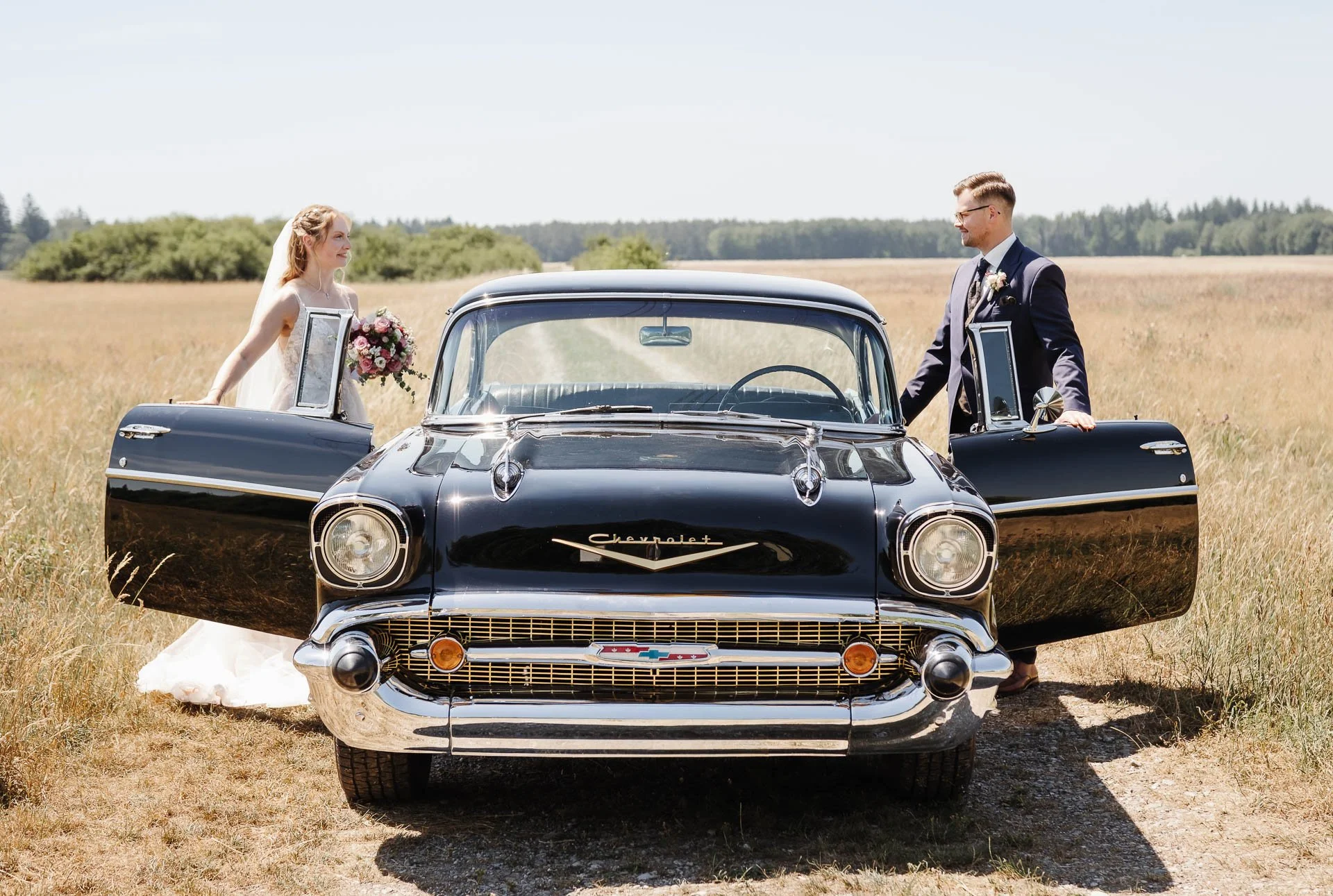 Das Bild zeigt einen schwarzen Oldtimer als Hochzeitsauto mit offenen Türen in einer sommerlichen Landschaft bei Augsburg, in den gerade die Braut und der Bräutigam einsteigen und sich über das Auto hinweg ansehen