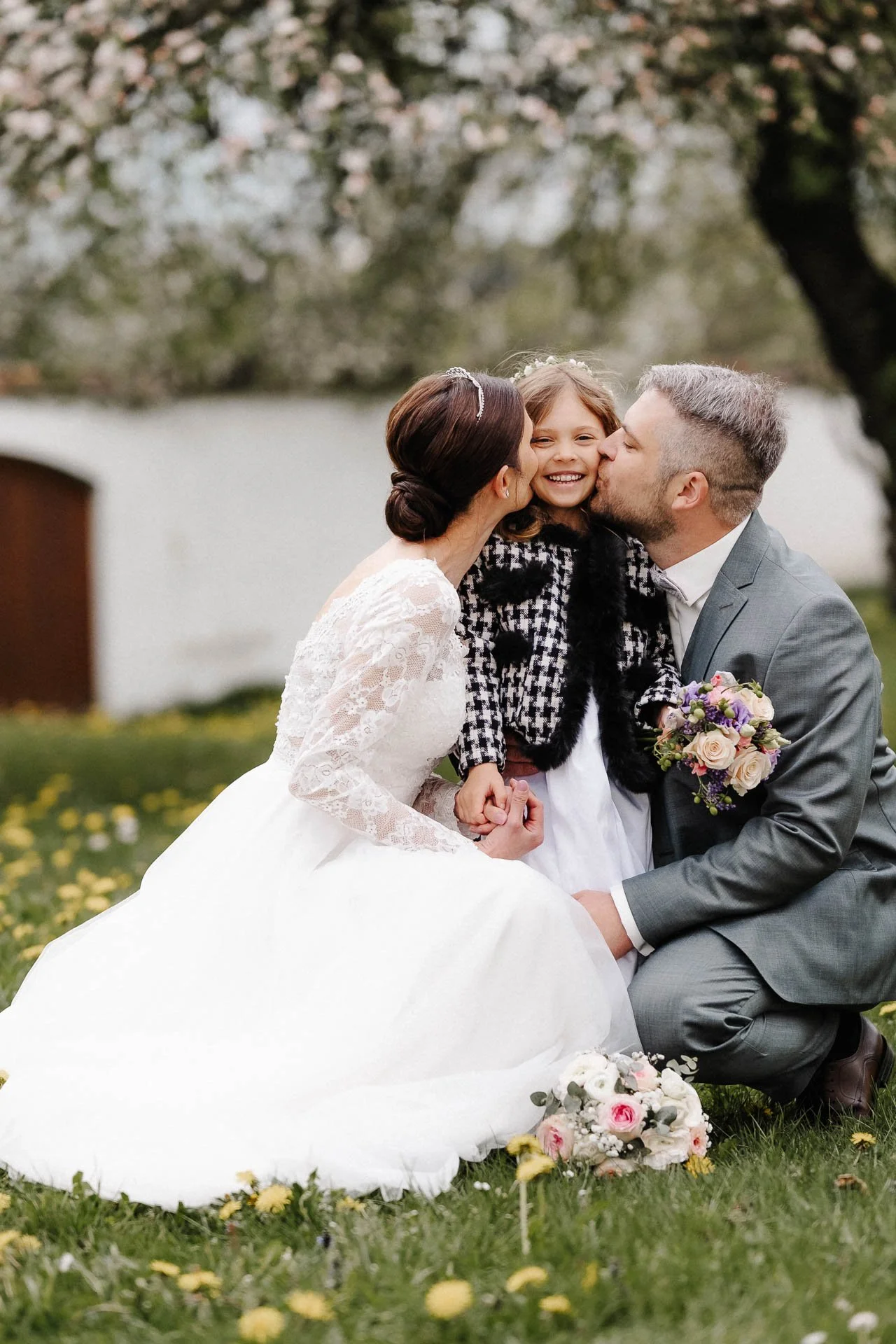 Hochzeitsfoto einer Familie mit Tochter bei Augsburg, bei dem die Eltern ihrem Kind von beiden Seiten einen Kuss auf die Wangen geben