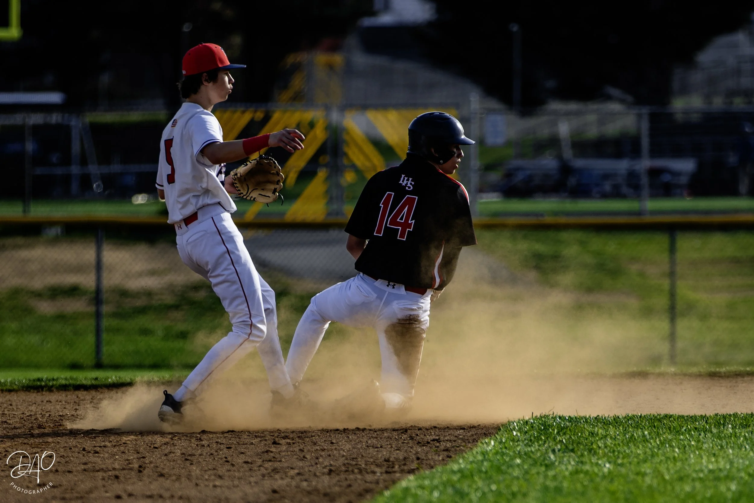 Two young boys playing baseball on a field, with one sliding into a base while the other is catching or preparing to catch a ball. The player in white is wearing a red cap and gloves, and the other player in black has a batting helmet.