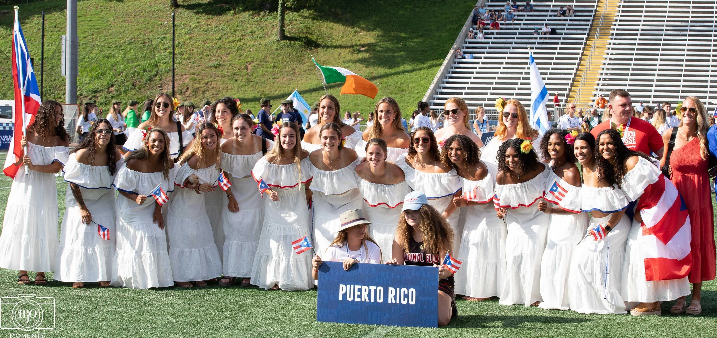 Team Photo. Group of women and children dressed in white dresses, holding Puerto Rican flags, at an outdoor event with spectators in bleachers and flags in the background.