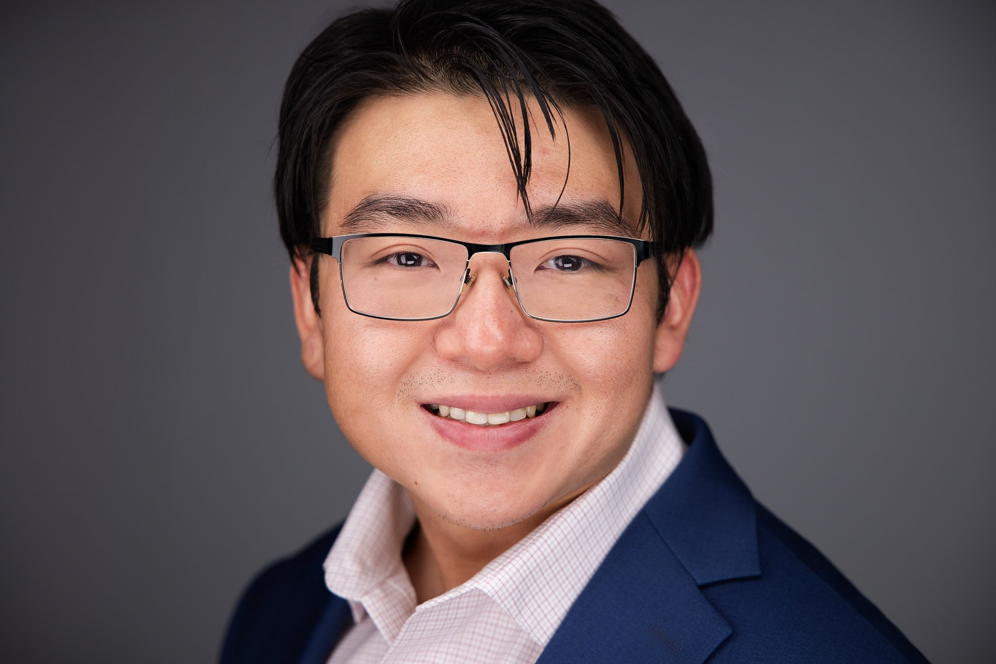 Headshot - Close-up portrait of a smiling man with black hair wearing glasses, a blue blazer, and a pink collared shirt against a gray background.