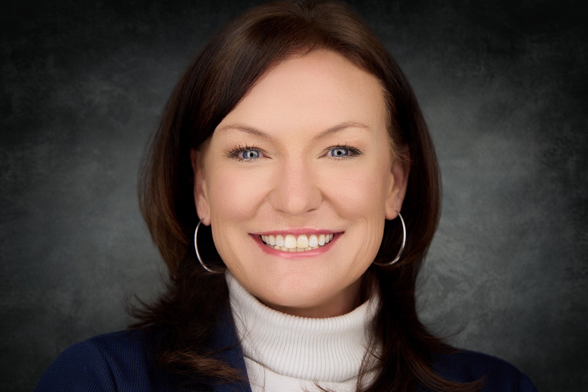 Close-up portrait of a woman smiling, with brown hair, blue eyes, earrings, wearing a white turtleneck and a dark blazer, against a dark gray background.