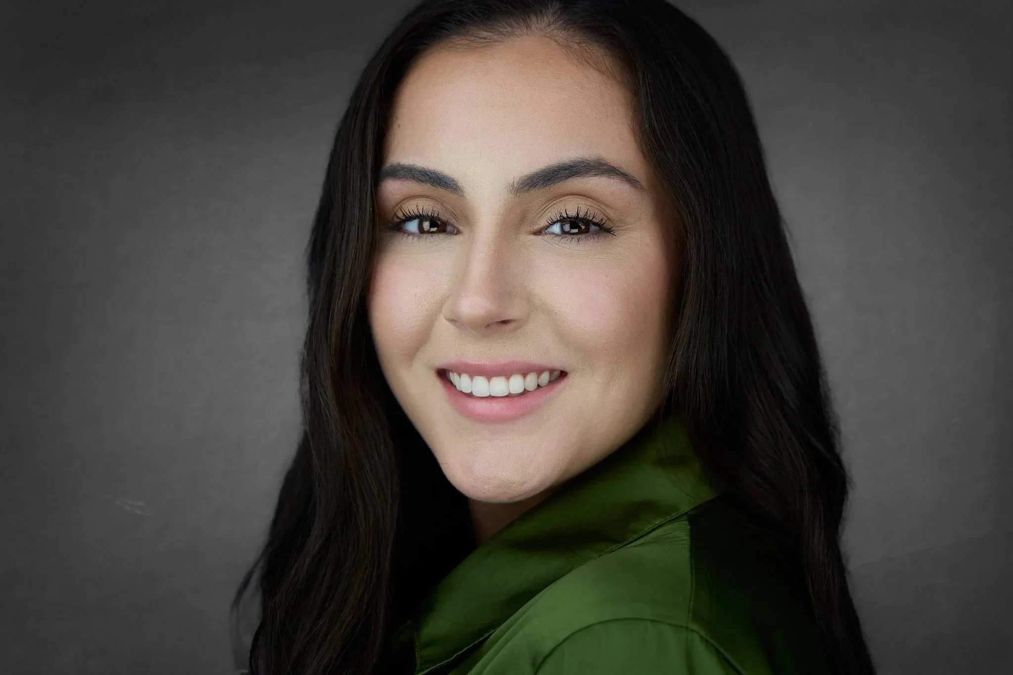A woman with long dark hair, brown eyes, and light skin smiling, wearing a green top, against a dark gray background.