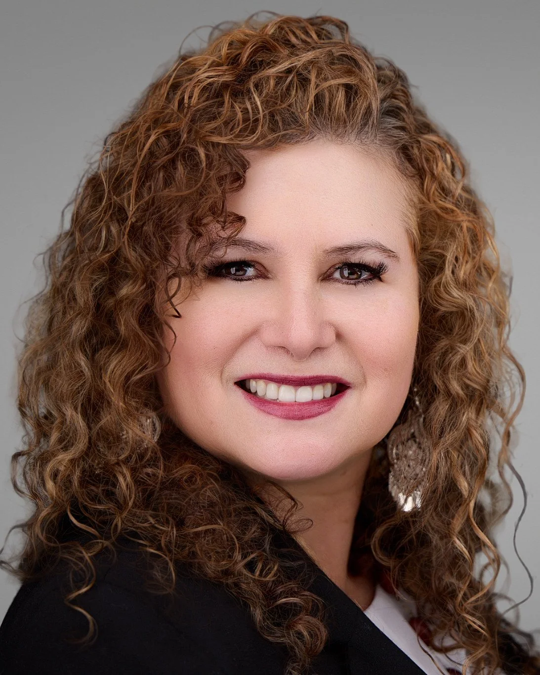 Portrait of a woman with curly red hair, smiling, wearing makeup, earrings, and a dark blazer.