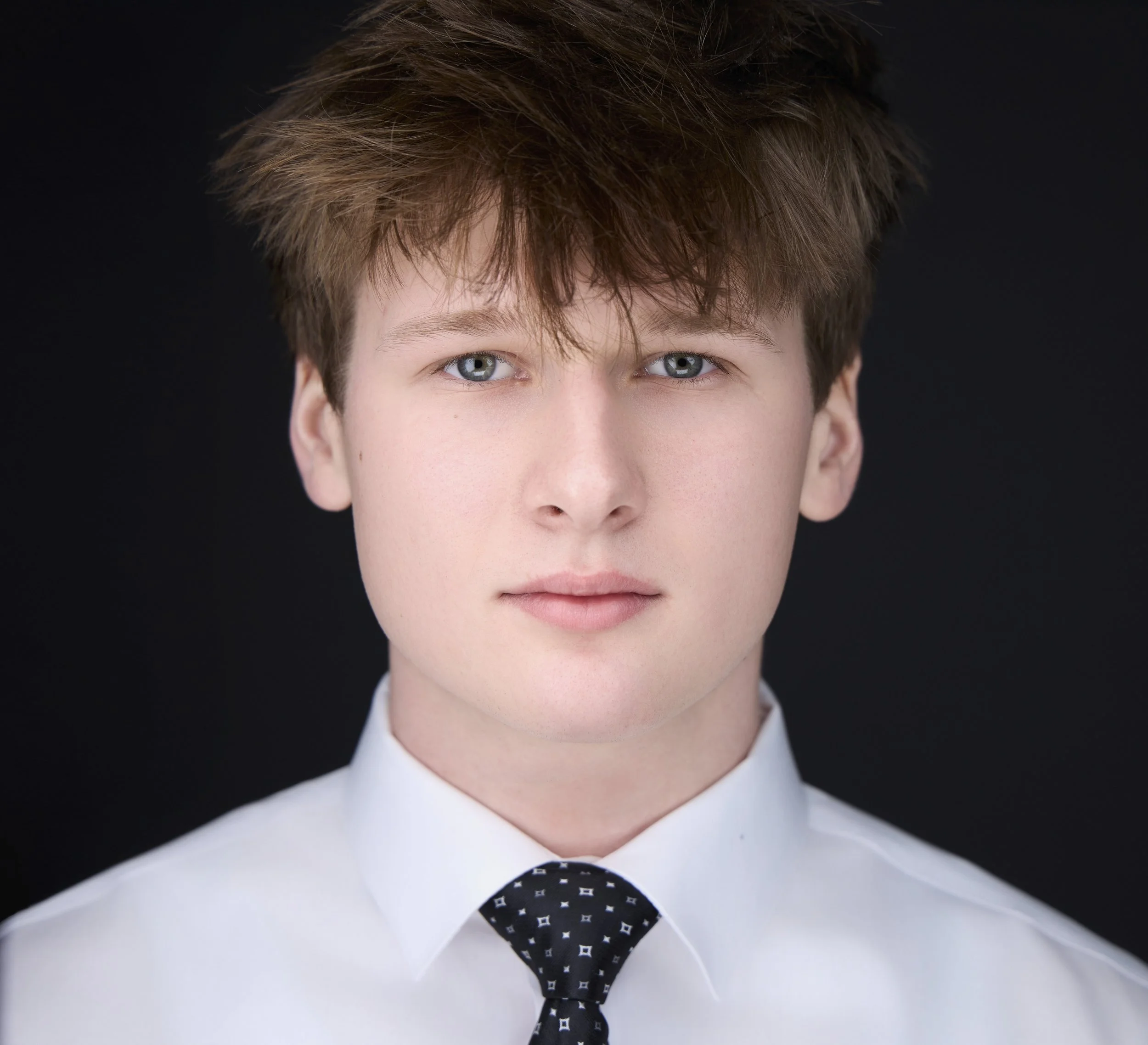 Headshot. Close-up of a person with blue eyes, light skin, and brown hair wearing a white shirt and a black tie with white geometric patterns, against a black background.