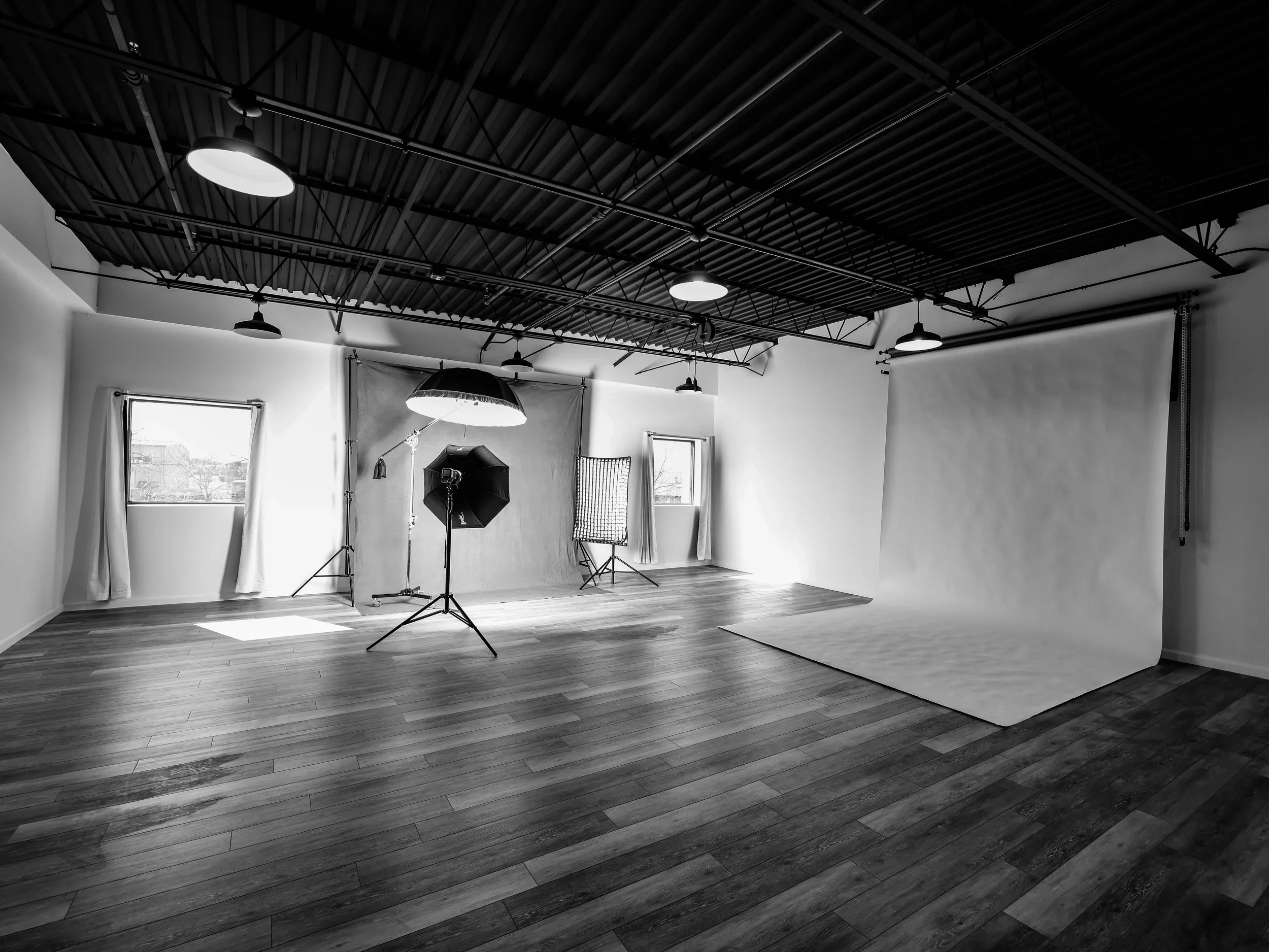 An indoor photography studioe. It features a studio setup with lighting equipment, backdrop, window, curtains, and wooden floor.