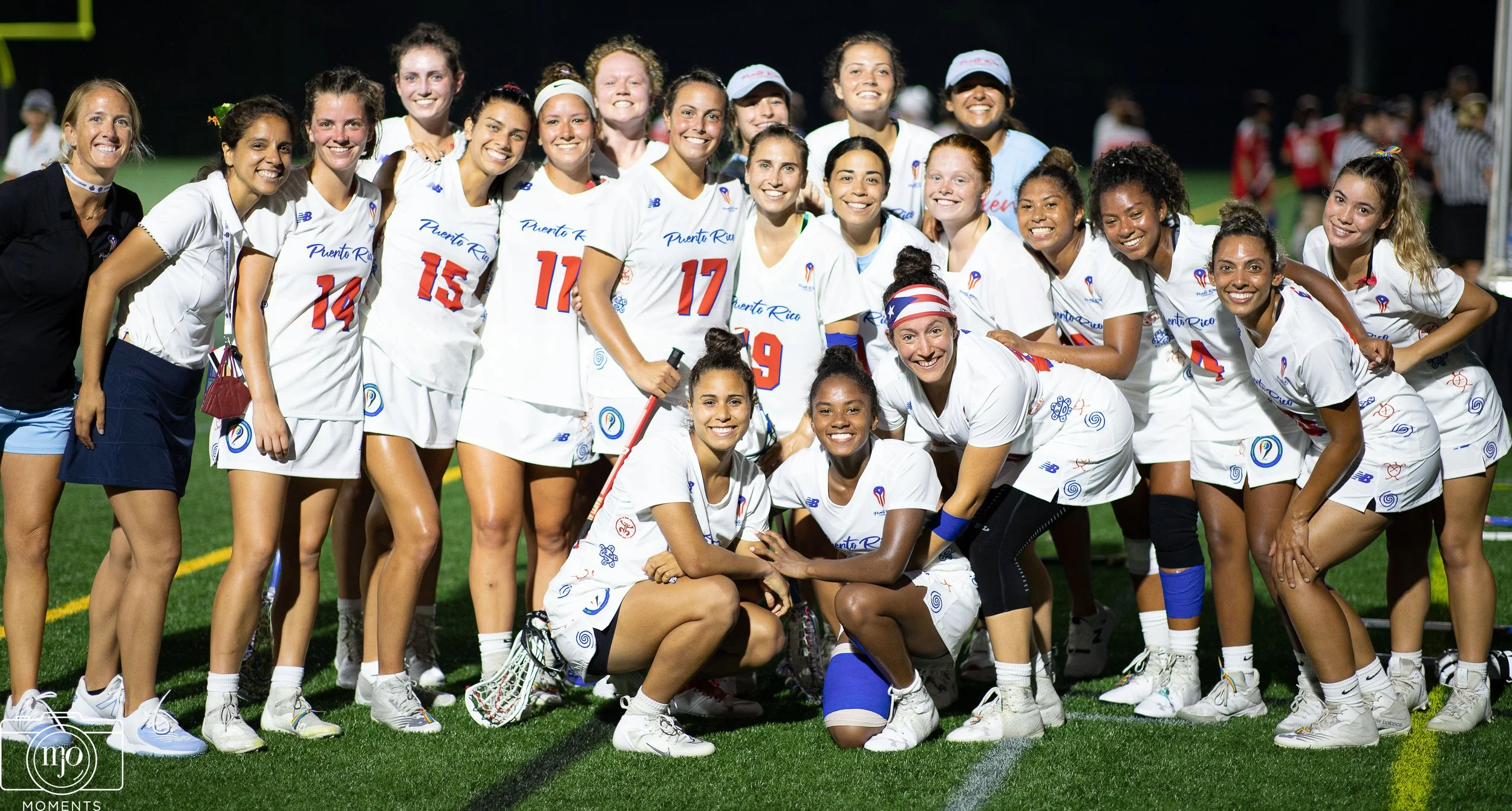 A group of women in white sports uniforms on a field, smiling and posing for the photo after a game, with some kneeling and others standing, under bright lights at night.