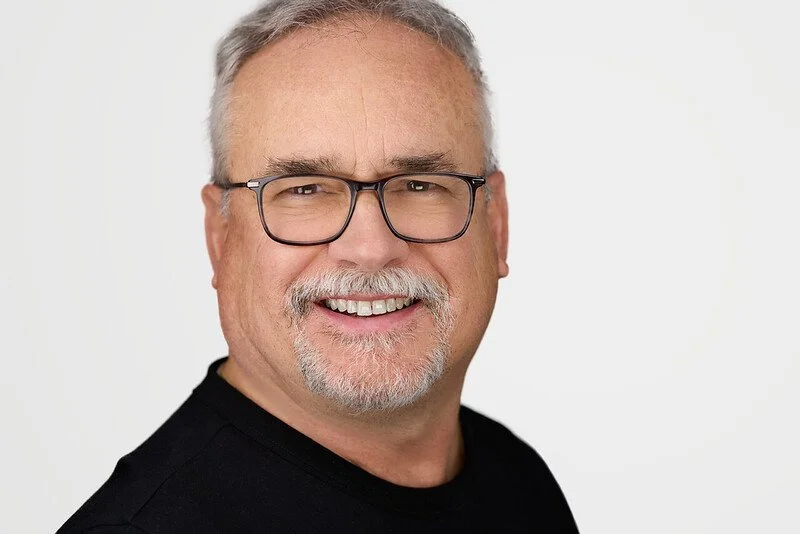 A smiling middle-aged man with gray hair, glasses, and a goatee, wearing a black shirt.