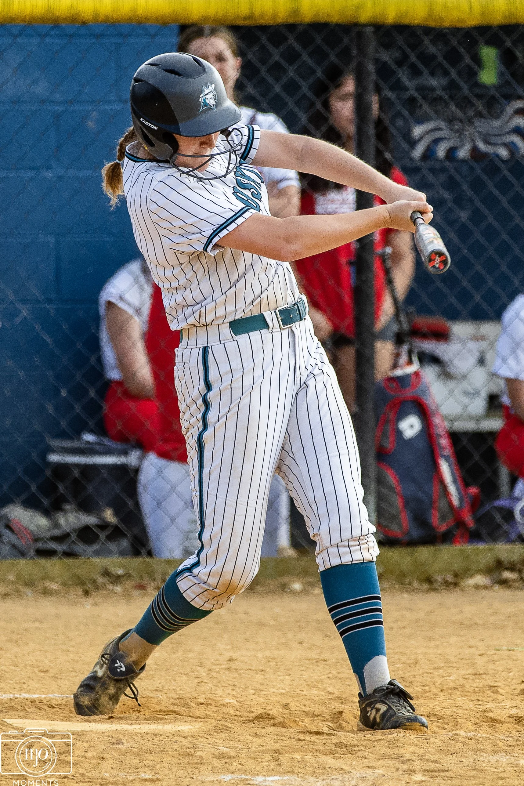A female softball player in striped uniform batting on a dirt field, wearing a helmet and mitt, with spectators and a chain-link fence in the background.