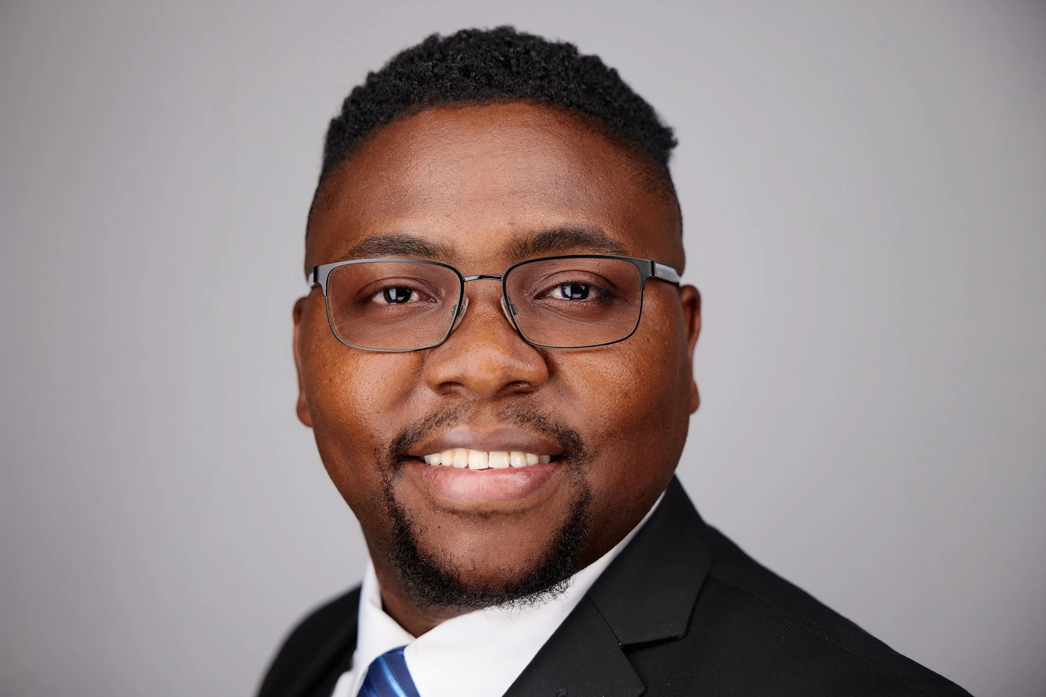 Headshot of a smiling man wearing glasses and a tuxedo with a blue tie, against a plain light gray background.