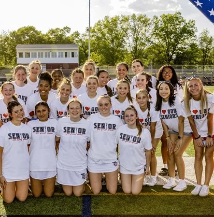 Team Picture. Group of young female track athletes in white shirts with 'SENIOR' writing, posing on a running track with green trees and a building in the background.