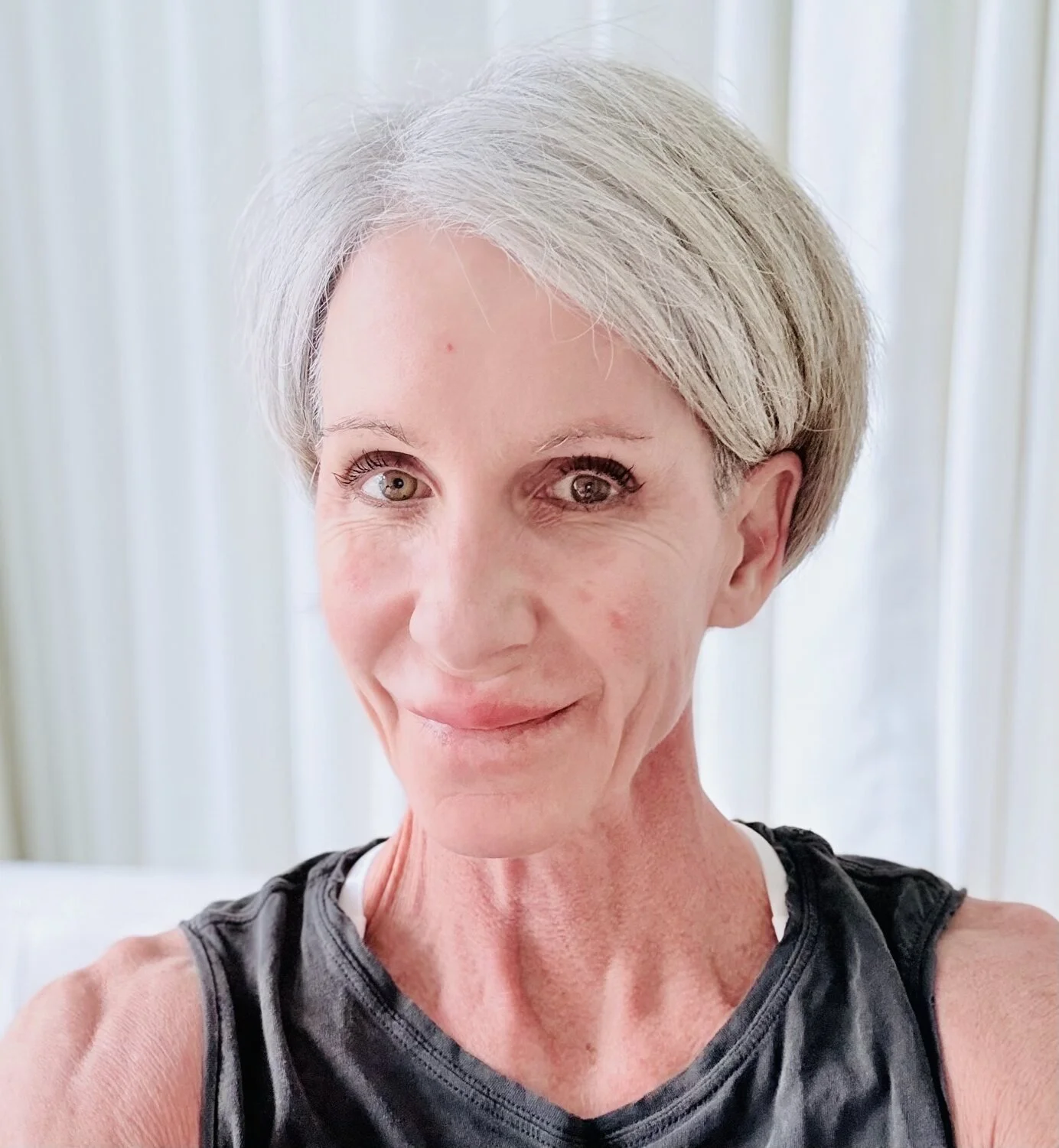 A middle-aged woman with short gray hair, smiling, wearing a black and white athletic top, in front of light-colored vertical blinds.