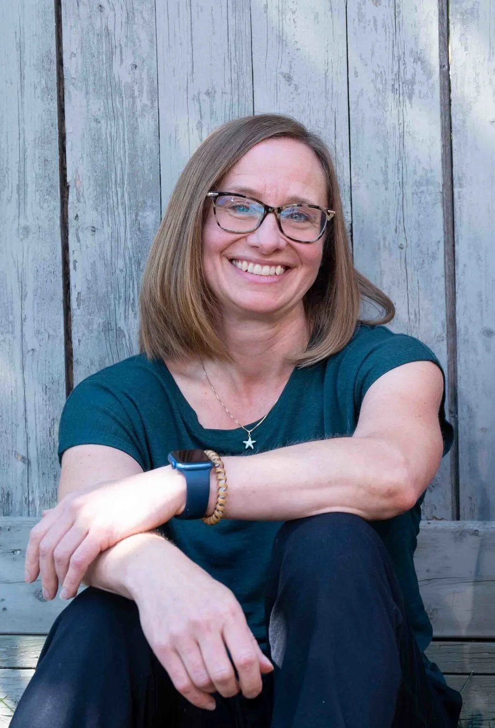 A woman with shoulder-length light brown hair, wearing glasses, a teal T-shirt, a star necklace, a smartwatch, and a wooden beaded bracelet, smiling while sitting outdoors against a wooden fence.