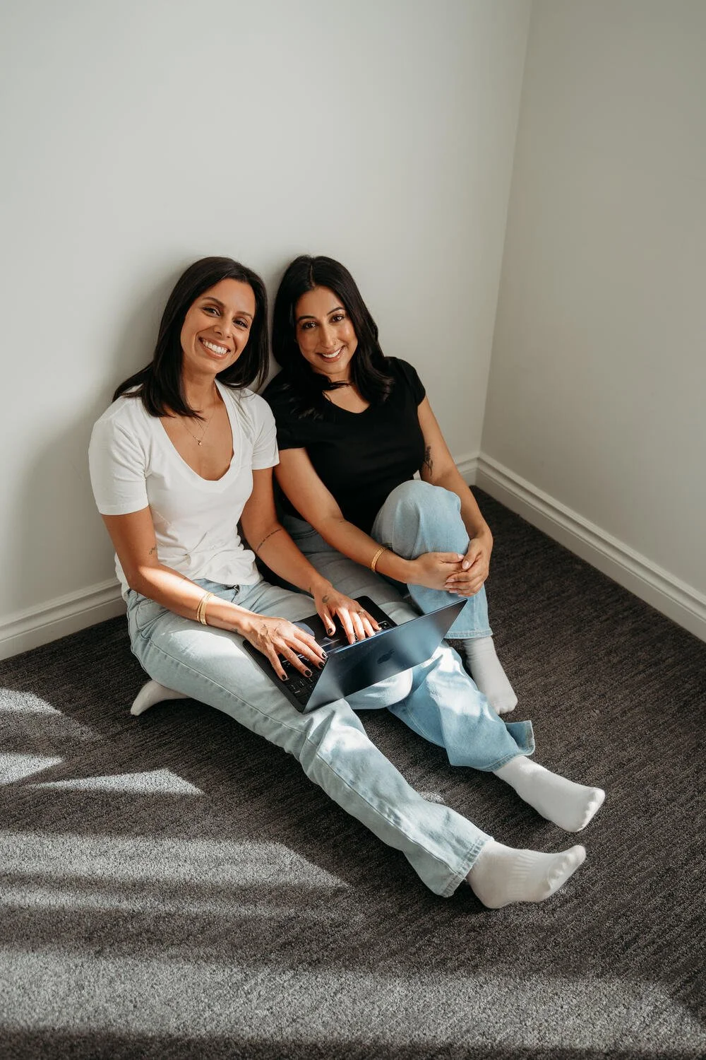 Two women sitting on the carpeted floor against a white wall, smiling and using a laptop, one in a white t-shirt and the other in a black t-shirt.