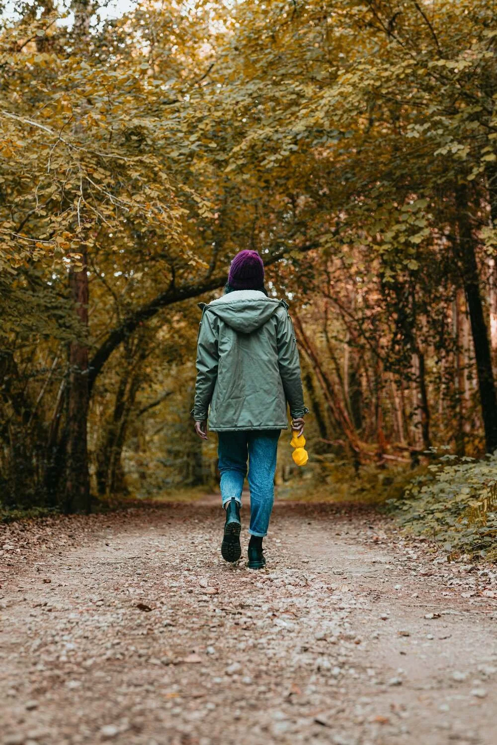 Person walking along a forest trail surrounded by fall foliage, wearing a green jacket, blue jeans, black boots, and a purple beanie, holding yellow items in their right hand.