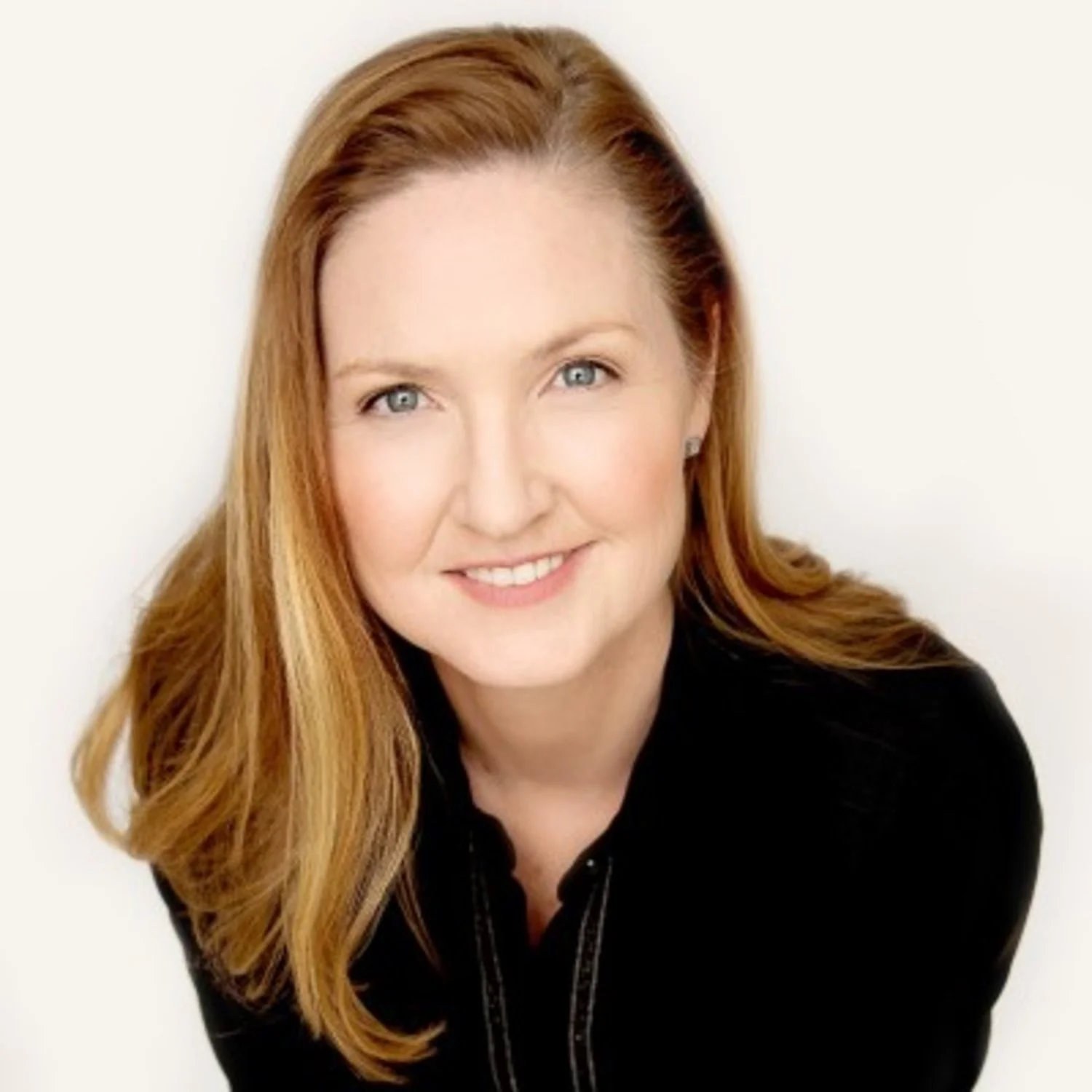 Close-up portrait of a woman with long red hair wearing a black top, smiling against a plain white background.