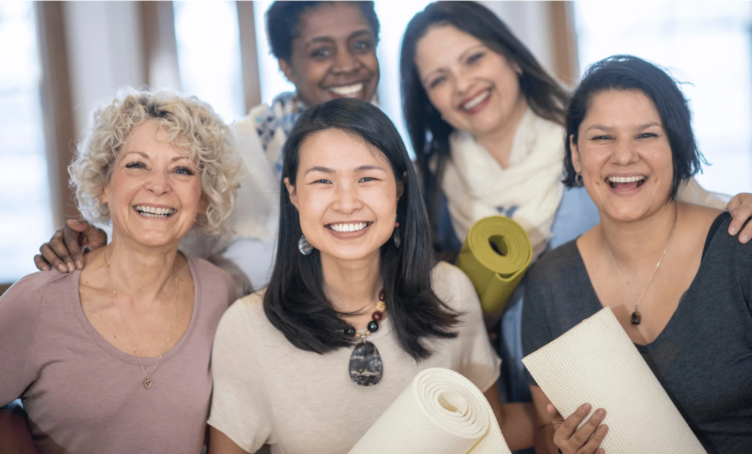 Five women smiling and holding yoga mats indoors with natural light.