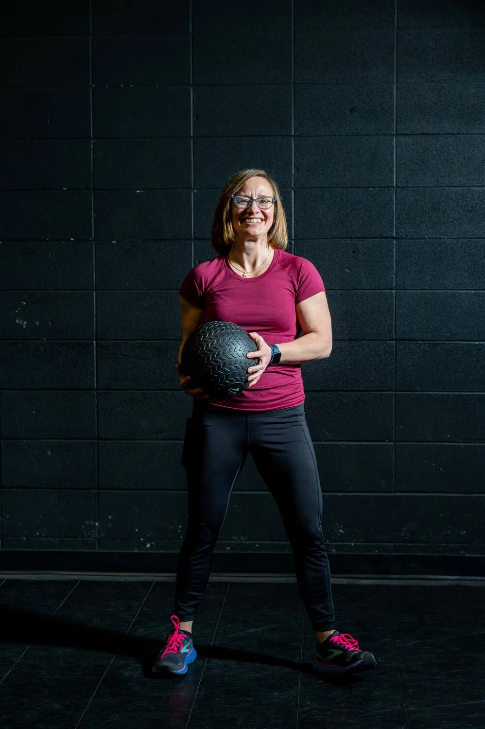 A woman in workout clothes holding a black textured medicine ball, standing in front of a black wall in a gym.