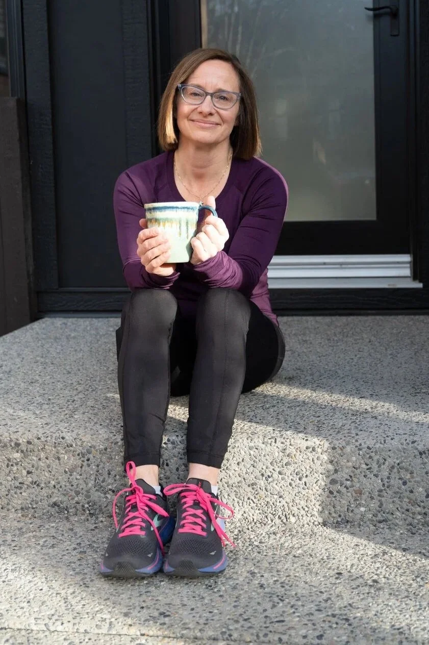 A woman sitting on a concrete step outside a building, holding a mug, smiling at the camera, wearing glasses, a purple long-sleeve shirt, black pants, and black athletic shoes with pink laces.