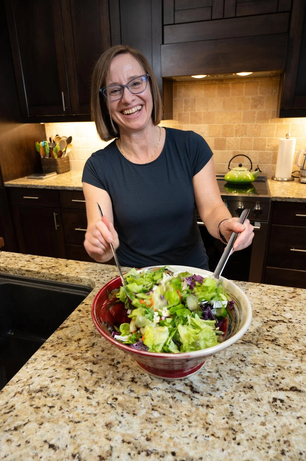 A woman with glasses and a black shirt smiling while tossing a salad in a bowl in a kitchen.