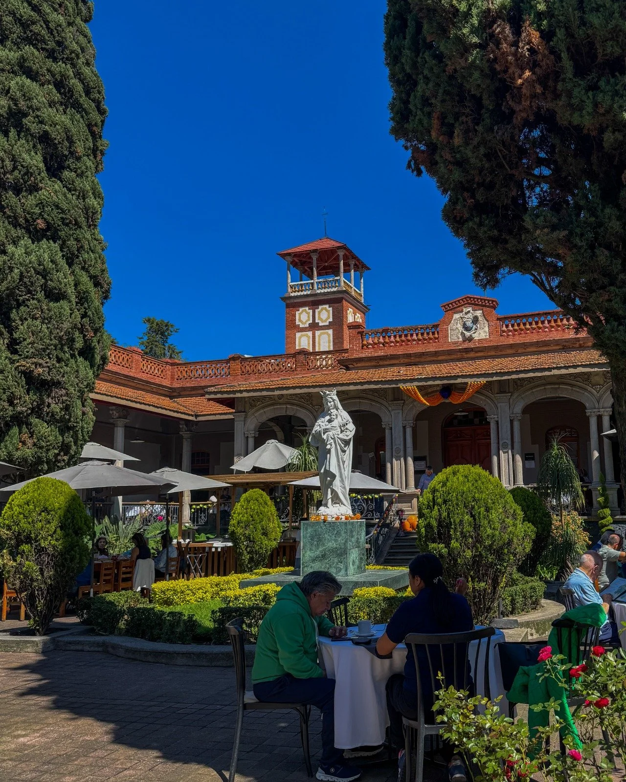 Escenario de una plaza en un edificio histórico con terraza y estatua de una figura religiosa en el centro. Personas sentadas en mesas al aire libre, rodeadas de árboles y plantas.