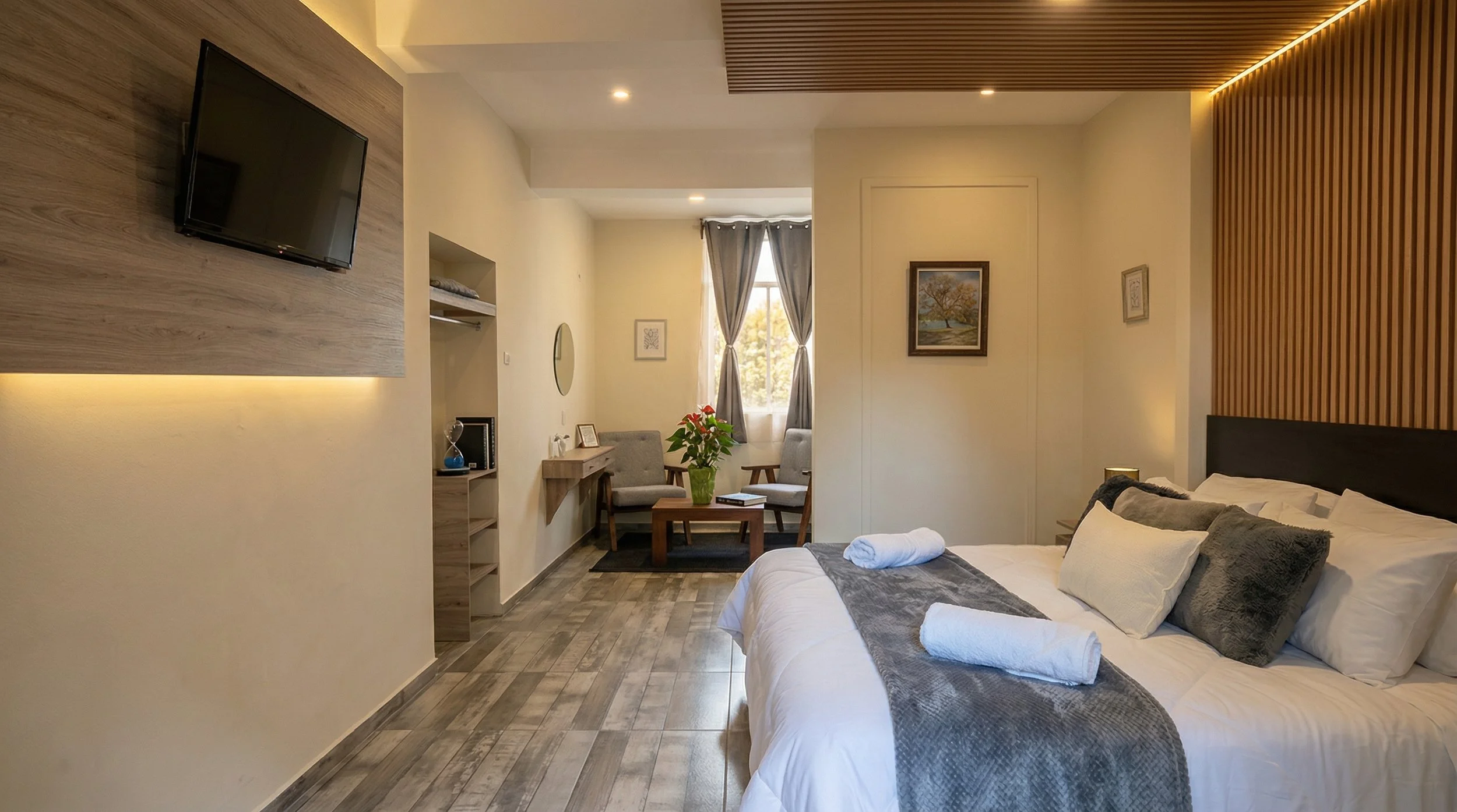 Habitación de hotel con una cama grande, cojines y toallas, área de descanso con dos sillones y mesa con planta, iluminación suave, paredes decoradas con cuadros y ventanas con cortinas grises, revestimiento de madera en el techo y paredes.