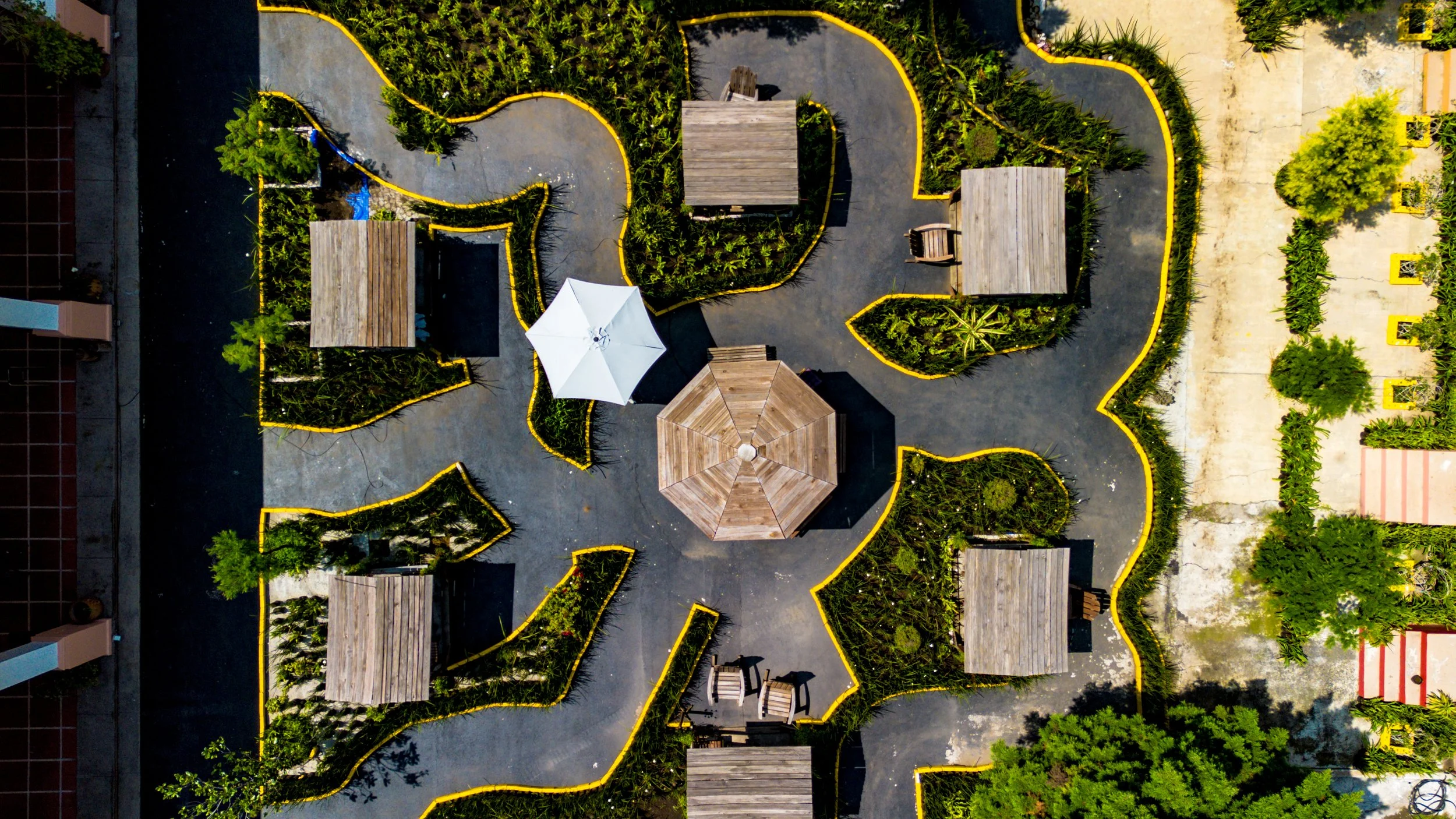 Vista aérea de un parque con senderos curvos, áreas verdes, varias pequeñas estructuras de madera, una sombrilla blanca, y asientos de madera.