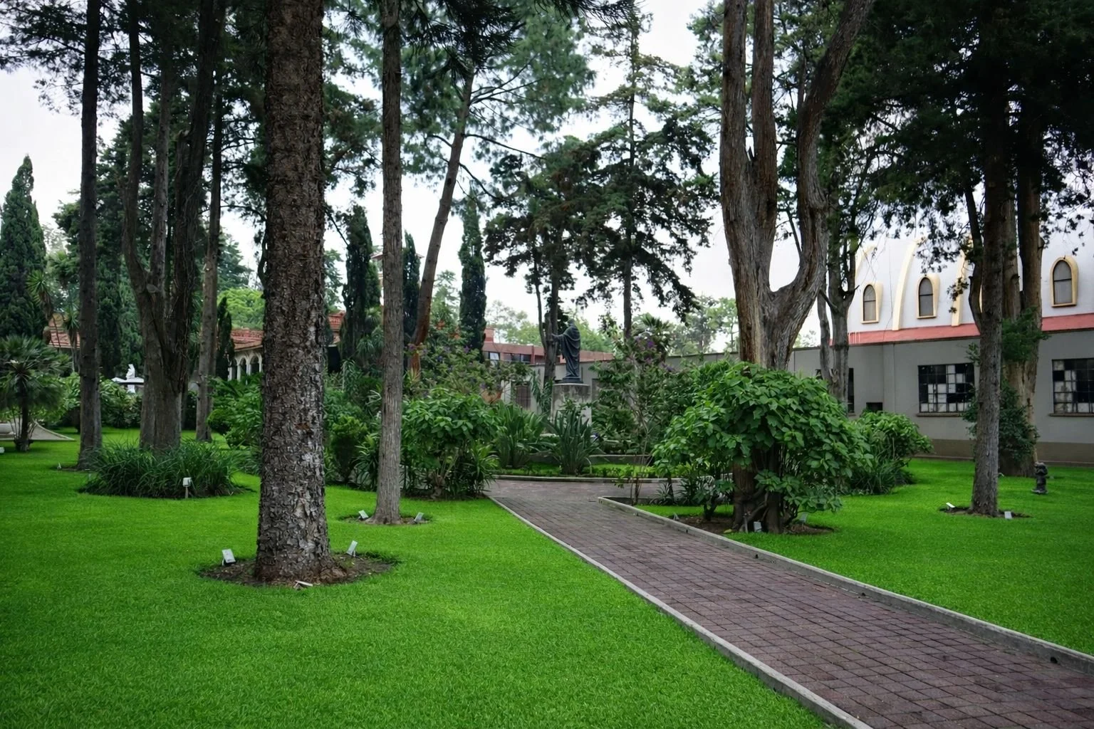 Jardín con árboles grandes, camino de ladrillos, plantas verdes y una estructura blanca en el fondo.