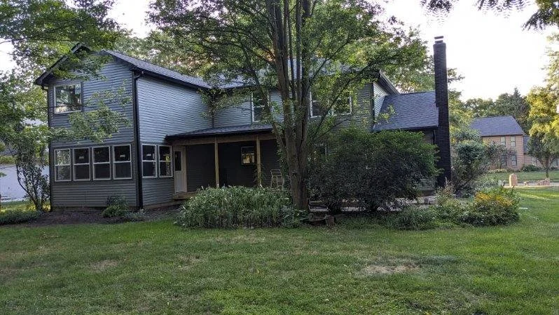 A two-story suburban house with gray siding, a black chimney, and a small porch, surrounded by a green lawn, bushes, and trees.