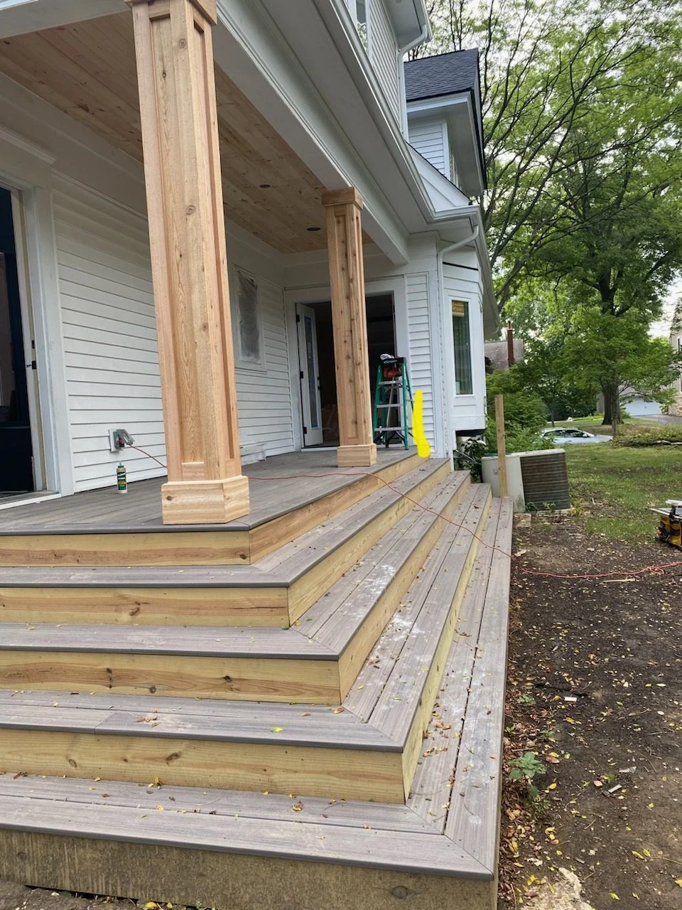 Construction of a wooden deck on the back of a house, with columns for a porch or balcony being built, surrounded by green trees and outdoor elements.