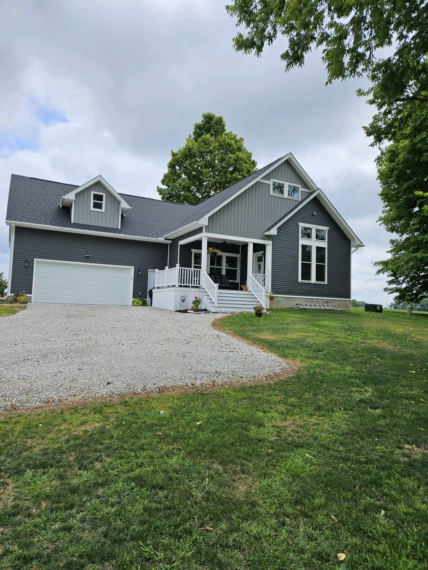A two-story house with gray and white exterior, attached garage, front porch with white railing, steps, and potted plants, surrounded by a gravel driveway and green lawn, with trees and a cloudy sky overhead.