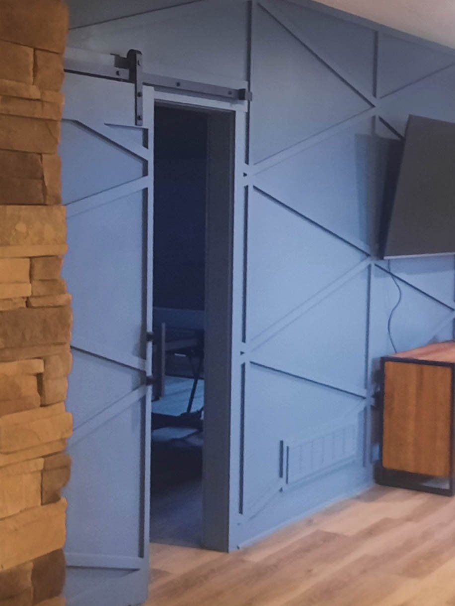 Blue sliding barn door with geometric panel design partially open, revealing a dark room with a chair inside, adjacent to a brick wall and a wooden cabinet.