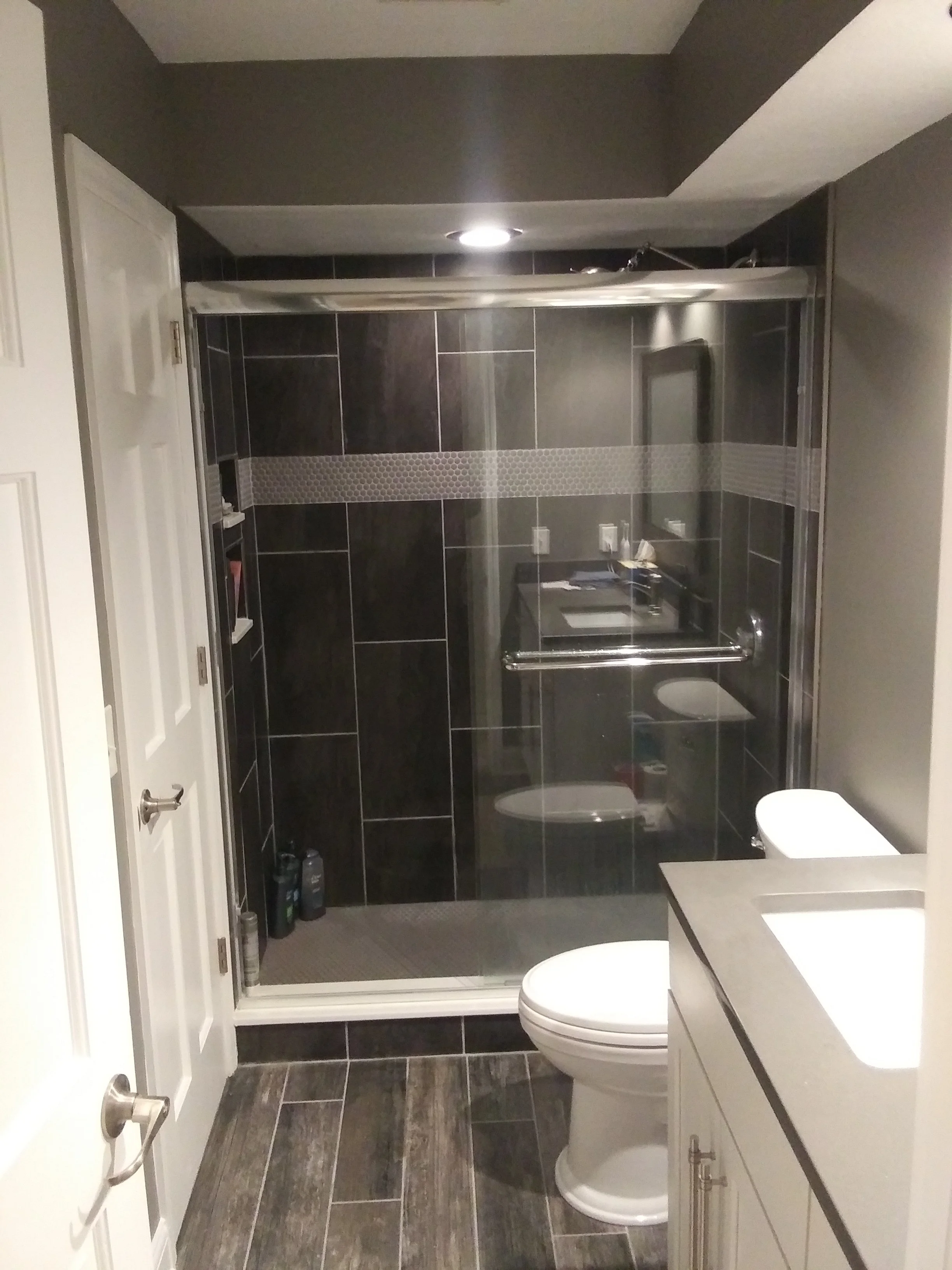 Modern bathroom with black tiled walls, a glass shower enclosure, a white toilet, and a white vanity with a countertop.