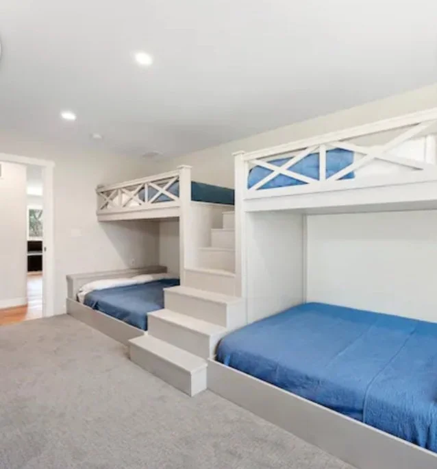 A bedroom with two sets of white bunk beds, each with a staircase and blue bedsheets, situated on a beige carpeted floor.