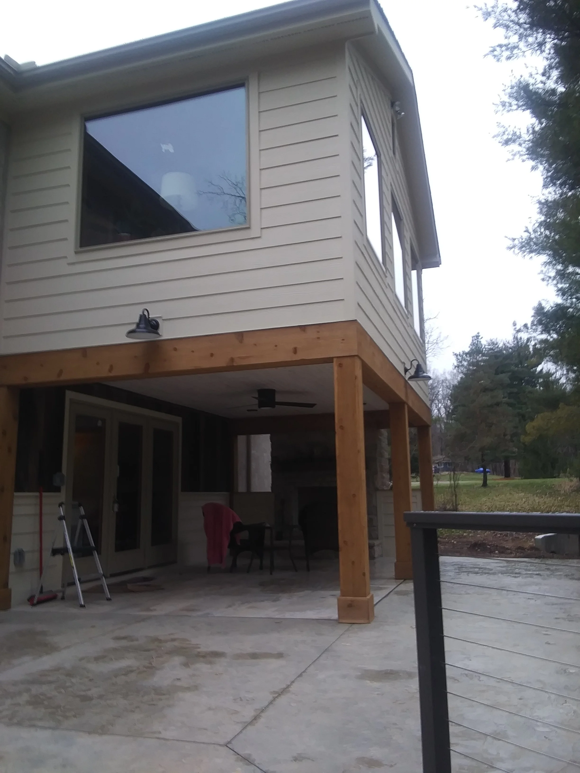 View of a two-story house with a deck, construction tools, and outdoor furniture, surrounding trees, and a cloudy sky.