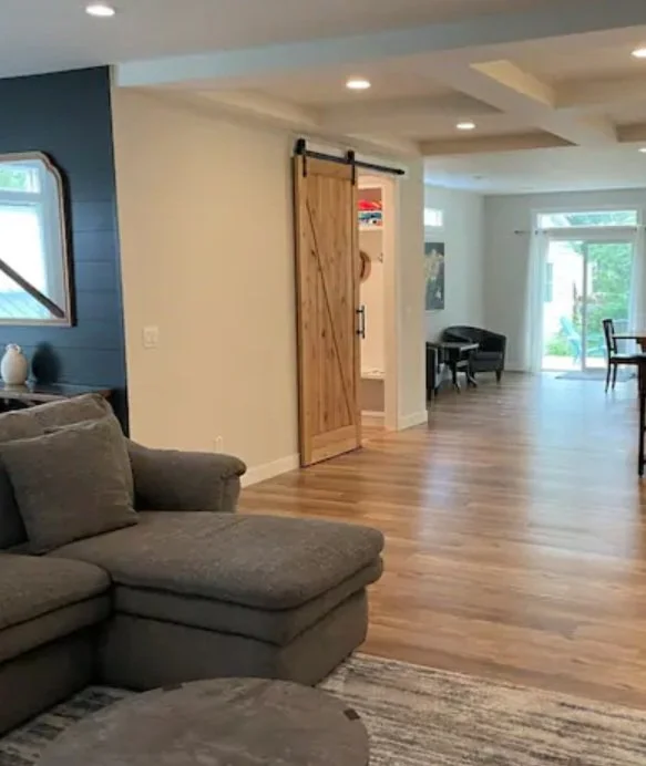 Living room with gray sectional sofa, wooden flooring, a sliding barn door, and a dining area with chairs near large windows.